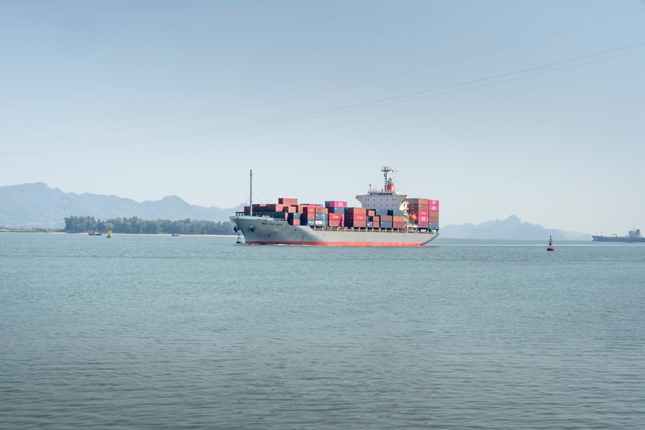 Exportação Agronegócio — A large container ship sails in calm waters under a clear sky, mountains in background.